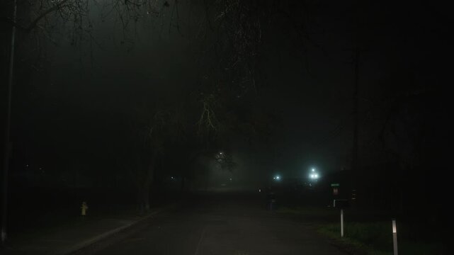 Moving backward along foggy street with approaching car moving in same direction and illuminating surroundings with bright headlights creating tense nighttime atmosphere with dense fog