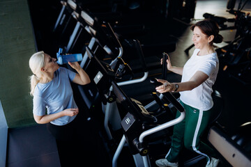 Fototapeta premium Two women engage in a lively gym session, one stands on an elliptical while the other enjoys a refreshing drink, encouraging each other in their fitness journey.