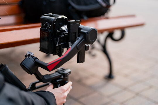 Professional camera on motorized gimbal held by operator near urban bench, illustrating modern videography equipment, social media production, and agile filming setup