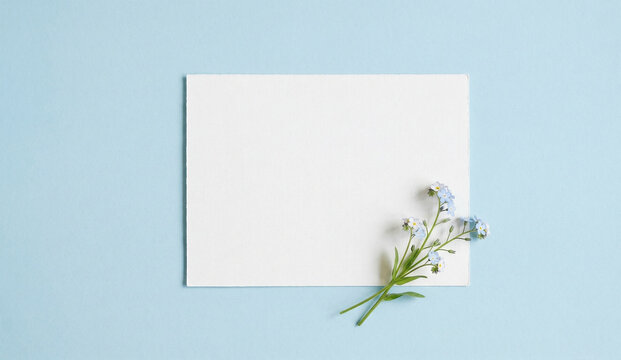 Empty white paper card with small blue flowers on a pastel blue background. Top view, flat lay, mockup for greeting card or wedding invitation.