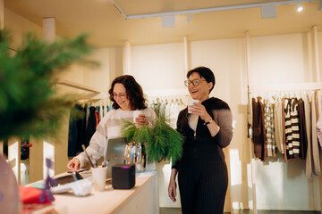 Friends share laughter and excitement in a stylish boutique. One arranges greenery while the other enjoys a warm drink. It is a delightful day of shopping and friendship.