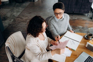 Two women are engaged in a lively discussion as they review important documents at a charming cafe. Notes and coffee surround them, creating an inspiring work atmosphere.