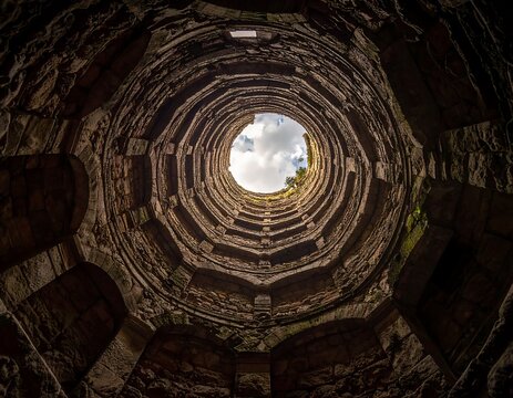 Ascending stonework spirals up a tall, aged well towards a bright opening with cloudy sky peering through
