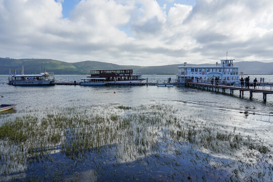 South Africa,Knysna Lagoon,Boats docked at a lakeside pier with people boarding, surrounded by calm water and distant hills under a cloudy sky.