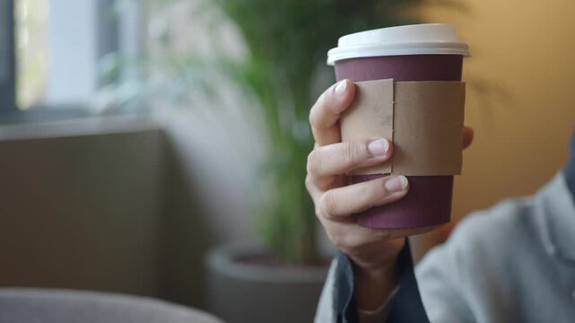 Woman holding disposable paper coffee cup with cardboard sleeve inside cafe interior clip.