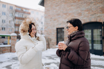 Fototapeta premium Two friends stand outside in a winter wonderland, wrapped in cozy coats. They share smiles and warm drinks while enjoying a chilly yet cheerful day together in the city.