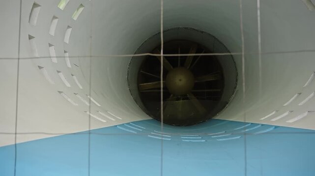 Close-up view inside a large industrial wind tunnel duct featuring an axial fan with prominent blades, a protective safety grid, and perforated curved walls used for aerodynamics testing and airflow