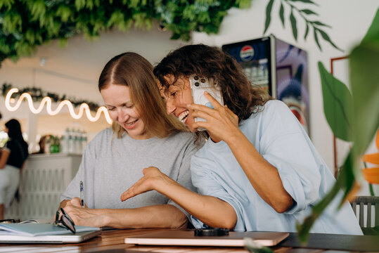 Two women sit at a table in a cafe. One writes in a notebook while the other talks on the phone and smiles. They enjoy their time together in a bright setting.