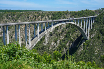 Fototapeta premium South Africa,Bloukrans Bridge,A modern concrete arch bridge spans a deep canyon with lush greenery and a vehicle crossing under a clear blue sky.