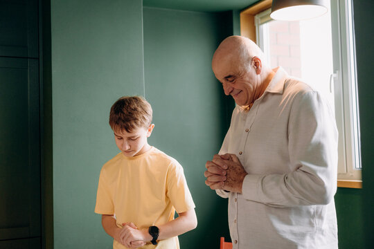 A man and a boy stand side by side with their hands together in prayer. They are inside a room with green walls and a window that lets in light. The time is likely morning.
