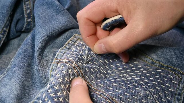 closeup shot of hands stitching a japanese Sashiko cross pattern onto the backpocket of a denim Blue Jeans.jujizashi