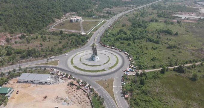 aerial turning view of the Preah Thong and Neang Neang statue at a major roundabout in Sihanoukville, Cambodia, featuring circular traffic flow, roadside markets, and lush greenery.