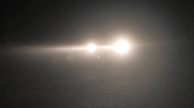 Car headlights cutting through dense fog at night, bright vehicle beam piercing thick mist on dark road creating dramatic low visibility driving atmosphere