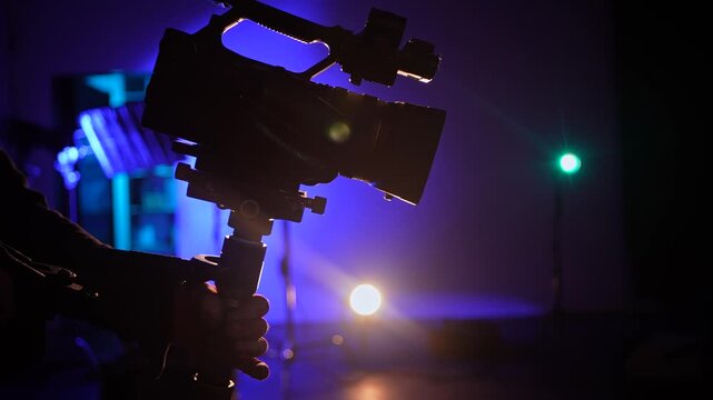Silhouette of a camera operator holding professional video equipment in a blue lit studio with glowing spotlights. Concept of filming, media production, broadcasting, backstage work and entertainment.