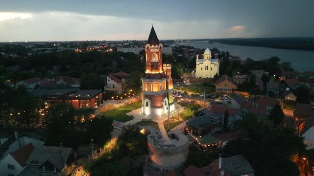 Dramatic aerial rotation of illuminated Gardos tower at night under a stormy sky with background lightning, Zemun, Belgrade, Serbia.