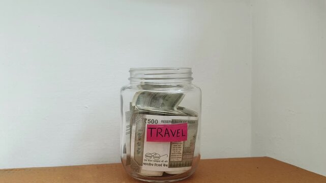 Close up of hand putting india rupee currency note inside transparent glass jar isolated on table with travel word or text label. Saving money for future dream vacation holiday or world tour concept.