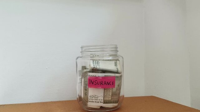 Close up of hand putting india rupee currency note inside transparent glass jar container isolated on table with Insurance word or text label. Saving money for next Insurance premium payment concept.