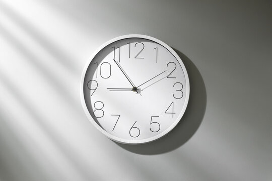 Minimalist white wall clock with black numerals on light gray wall, dramatic sunlight casting long striped shadows, modern time concept.