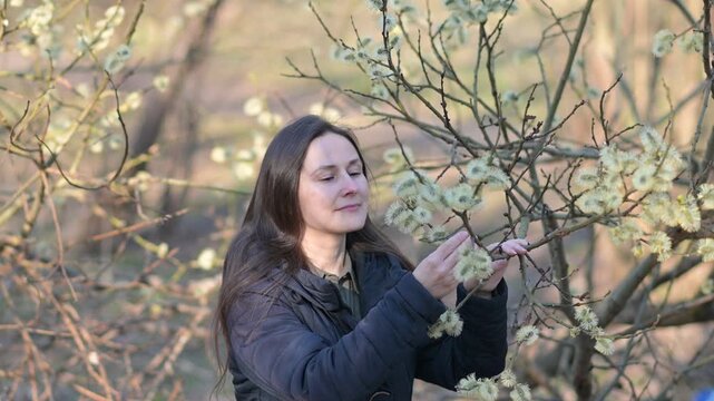 medium shot of a woman sneezing while standing close to flowering willow catkins in a spring forest. The scene clearly illustrates seasonal allergic rhinitis, hay fever symptoms