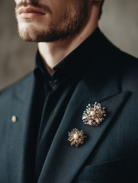 Man in tailored dark navy suit wearing gold, silver, and pearl brooches on collar against muted gray wall, luxury menswear fashion portrait