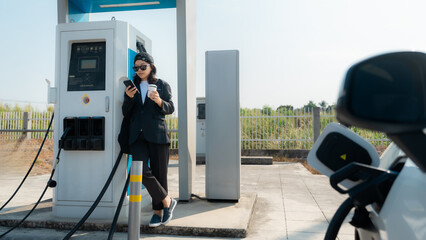 Young woman travel with EV electric car charging in green sustainable city outdoor garden in summer. © 2B