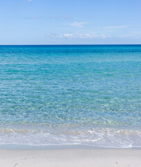 Fototapeta premium Turquoise sea waves gently lapping sandy beach shoreline in Sardinia, Italy. Calm Mediterranean waters under clear sky. Coastal summer landscape with pristine water.