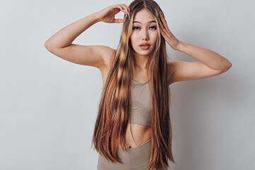 Fototapeta premium Young woman with long straight hair and casual top posing with hands near head against plain background, fashion and beauty concept with natural makeup.