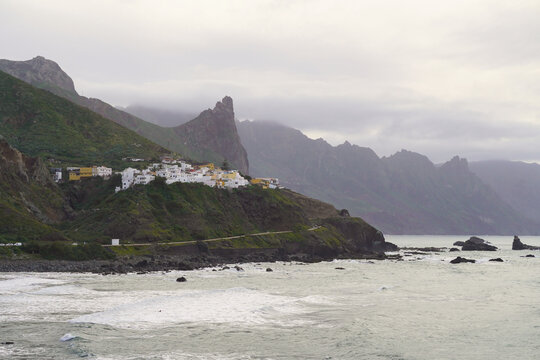 The coastal village of Almaciga nestled on a steep green hillside in Anaga, Tenerife. The image shows traditional white houses overlooking the rugged Atlantic shore with crashing waves.