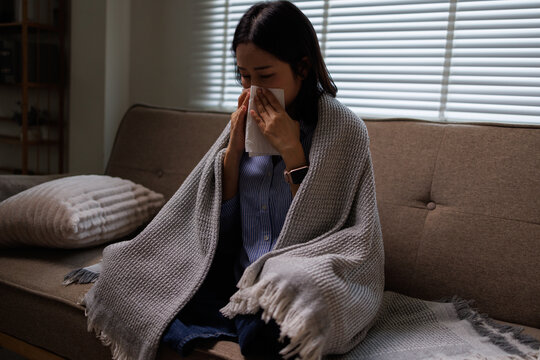 Woman feeling unwell and shivering under a blanket, resting on a sofa while blowing her nose with a tissue due to a cold or flu, experiencing illness symptoms indoors