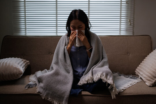 Young woman on a sofa wrapped in a blanket, sneezing into a tissue while sick at home, resting with cold or flu symptoms and seeking comfort during illness and isolation
