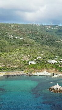 Scenic coastline of Centuri with turquoise water, hillside houses, and wind turbines on the ridge under a cloudy sky. A harmonious blend of nature and renewable energy.