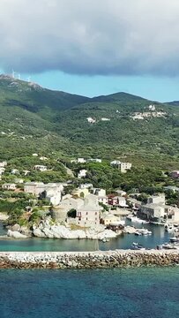 Aerial view of the picturesque coastal village and harbor of Centuri, Corsica, surrounded by lush green hills and clear blue water under a partly cloudy sky.