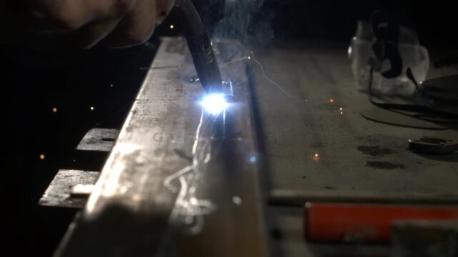 A person is welding metal in a workshop. Sparks fly as the weld joins two pieces of metal. The setting is dark, and the light from the welding creates bright flashes.