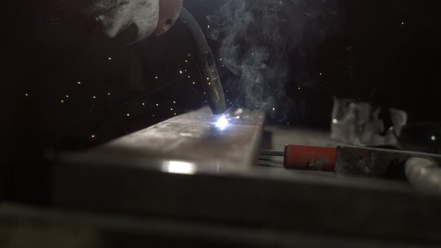 A person works on a metal piece in a workshop after dark. Sparks fly as welding happens, creating bright lights in the room. Tools and metal parts are visible in the background.