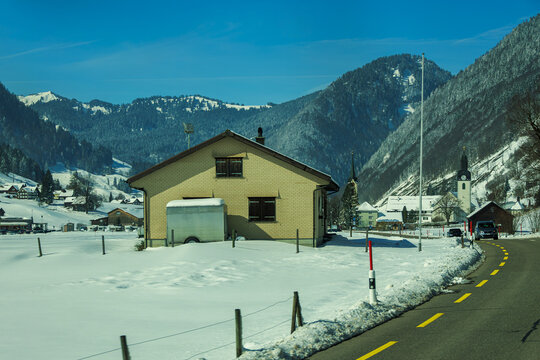 Picturesque winter scene featuring snow covered landscape with house and winding road leading through mountainous area. clear blue sky enhances serene atmosphere, while distant church adds touch