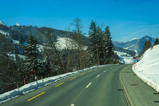 Scenic winter road curves through snowy mountainous landscape, bordered by tall evergreen trees under clear blue sky. serene setting evokes sense of tranquility and adventure