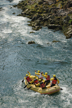 A group of rafters in a yellow boat wearing helmets and life jackets paddling through white water rapids on a rocky river
