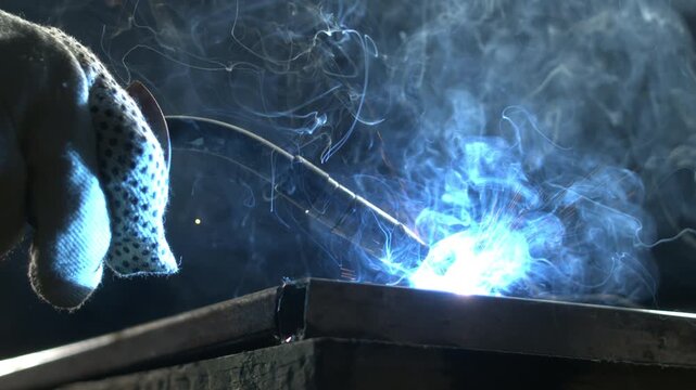 A person is welding metal in a workshop at night. Sparks fly as the welding torch creates bright light. The setting is dark, highlighting the work being done.