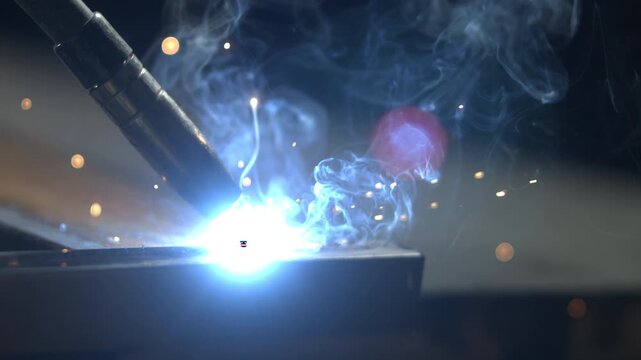 A person is welding metal pieces together in a workshop. Bright sparks are flying as they work, focusing on the task. Equipment and tools are visible in the background.