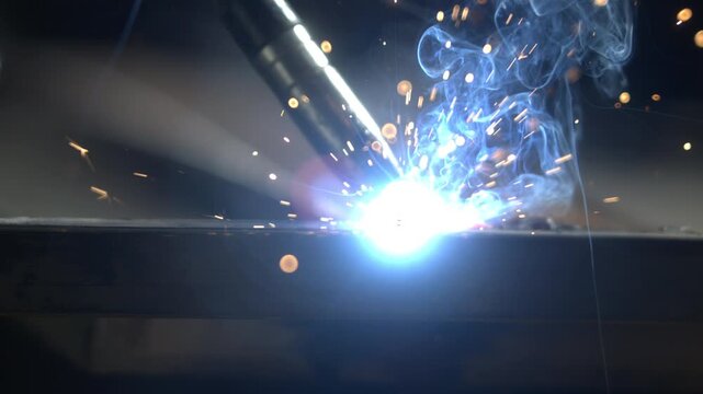 A welder works on metal at a workshop. Sparks and smoke arise as the torch cuts into the material. Tools are used to shape and join pieces of metal.