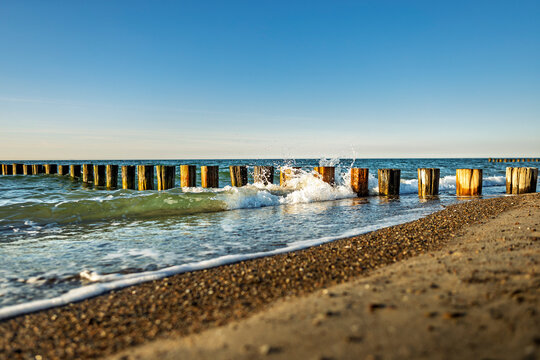 Diagonal shoreline with groynes acting as breakwaters, waves crashing and freezing into droplets and spray against a clear blue cloudless sky, dynamic coastal seascape motion scene