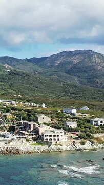 A scenic coastal village with traditional buildings nestled by the sea and backed by lush green mountains in Centuri, Corsica. Clear blue water and dramatic landscape create a tranquil Mediterranean