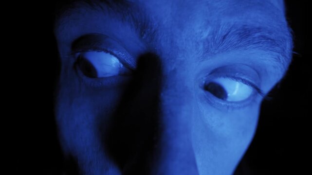 Male face stares into camera with wide terrified eyes under monochromatic blue light. Extreme close up captures rapid blinking and intense paranoid expression in darkness.