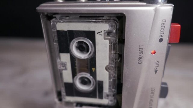 A silver microcassette recorder operates with the tape spinning and the red record light illuminated. Concept of retro technology, journalism, surveillance, and preserving analog audio history.
