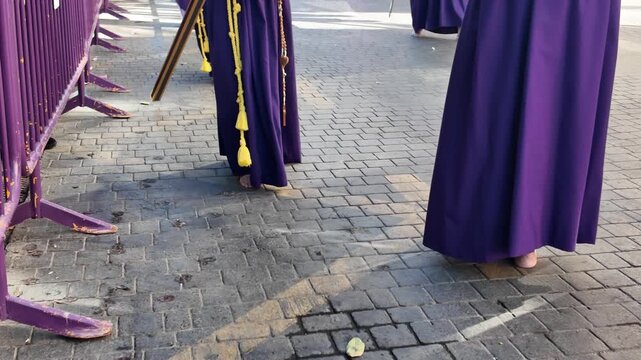 Nazarenos penitents barefoot wearing purple robes and pointed hoods walking on cobblestone street during Semana Santa Holy Week Catholic procession Spain traditional religious event