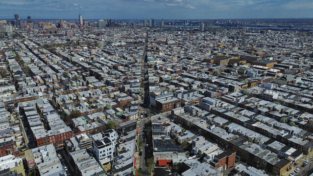 Expanding residential field near S 10th St, Tasker St reveals uniform blocks transitioning toward taller Center City structures. Flat roofs, repeating masonry, soft atmospheric light, Philly, PA