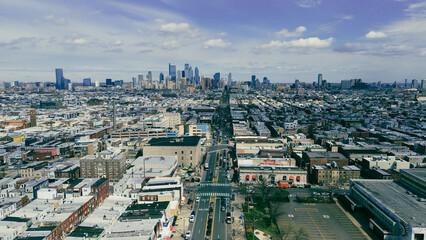 Obraz premium Central corridor ascent along Broad St reveals dense South Philly blocks converging toward the Center City skyline. Reflective roofs, brick gradients, soft atmospheric haze settle over Philly, PA