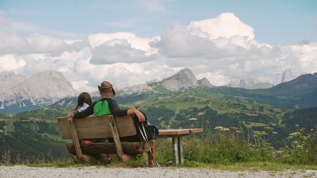 Caucasian couple hikers sit on a wooden bench, admiring the majestic Dolomites mountain range in Italy. A peaceful moment of adventure, wellness, and scenic trekking in South Tyrol.