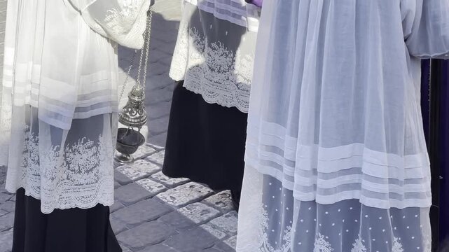 Young altar servers acolytes dressed in white lace robes and black vestments standing in sunlit outdoor religious ceremony traditional Christian Catholic procession setting