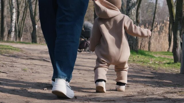 Toddler in a beige romper walks along a dirt trail in a park while an adult in jeans and white sneakers stays close. Concept of early childhood development and rhythmic outdoor exploration.